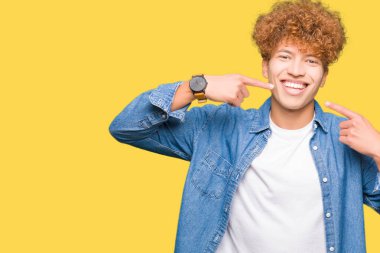 Kendinden emin gösterilen ve işaret parmakları diş ve ağız ile gülümseyen denim ceket giyen afro saçlı Genç yakışıklı adam. Sağlık kavramı.