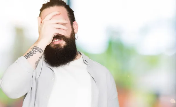 Young man with long hair, beard and earrings smiling and laughing with hand on face covering eyes for surprise. Blind concept.