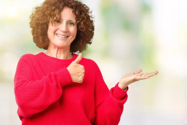 Güzel orta ager kadın kıdemli kırmızı kış kazak üzerinde arka plan gösteren palm el izole ve mutlu ve neşeli gülümseyen thumbs up, ok hareketi ile yapıyor