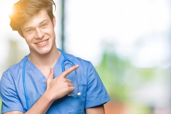 年轻的医生穿着医疗制服在孤立的背景愉快 脸上的微笑用手和手指指着一边 脸上带着快乐和自然的表情 — 图库照片