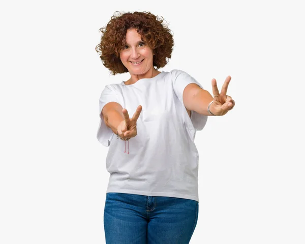 勝利のサインをやって指を表示するカメラを笑みを浮かべて孤立の背景に白い シャツを着て美しい中間エイガー年配の女性 — ストック写真