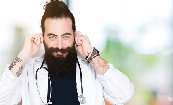 Doctor with long hair wearing medical coat and stethoscope covering ears with fingers with annoyed expression for the noise of loud music. Deaf concept.