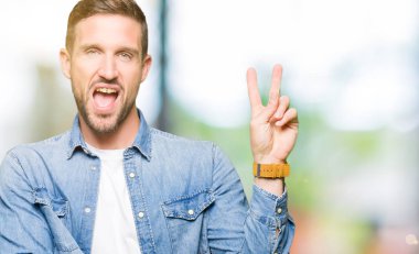 Mavi gözlü ve sakallı, kot ceket giyen yakışıklı adam mutlu yüzle gülümseyerek zafer işareti yaparak kameraya göz kırpıyor. İki numara.