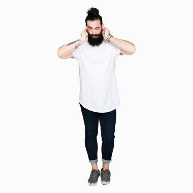 Uzun saç ve sakal casual beyaz t-shirt kapsayan kulakları parmakları ile yüksek sesle müzik gürültü rahatsız ifade ile giyen genç hipster adam. Sağır kavramı.