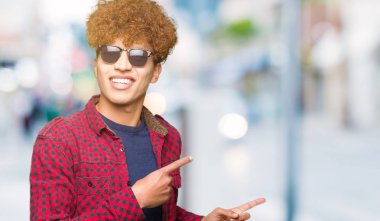 Gülümseyen ve iki elleri ve parmakları ile tarafına işaret kamera bakarak güneş gözlüğü takıyor afro saçlı Genç yakışıklı adam.