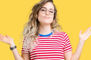 İzole arka plan clueless ve şaşkın bir ifadeyle kollar ve eller kaldırdı üzerinde gözlük güzel sarışın genç kadın. Şüphe kavramı.