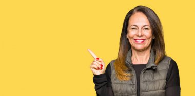 Yüzünde, kocaman bir gülümseme ile kış yelek giyen güzel orta yaş kadın kameraya bakarak yan el ve parmak ile işaret.