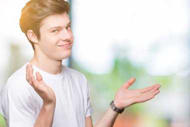 Üzerinde rahat beyaz t-shirt giyen genç yakışıklı adam arka plan clueless ve şaşkın bir ifadeyle kollar ve eller kaldırdı izole. Şüphe kavramı.