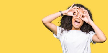 Kıvırcık saçlı Genç güzel kız rahat beyaz t-shirt giyerek dil yapışan dürbün gibi ok jest yapıyor, gözleri parmaklarla bakıyor. Çılgın ifade.