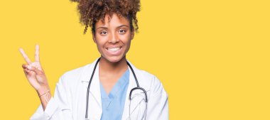 Genç Afro-Amerikan doktor kadın mutlu yüz zafer işareti yaparken kameraya göz kırpıyor ile gülümseyen izole arka plan yüzünden. İki numaralı.