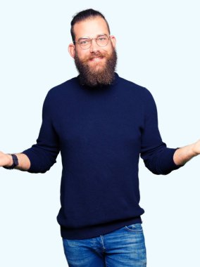 Gözlük ve balıkçı yaka kazak giyen genç sarışın adam her iki el açık avuç gösteren gülümseyen, sunma ve reklam karşılaştırma ve denge