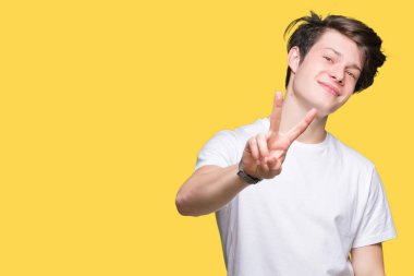 Zafer işareti yapıyor parmak gösterilen kamera seyir gülümseyen izole arka plan üzerinde rahat beyaz t-shirt giyen genç yakışıklı adam. İki numaralı.