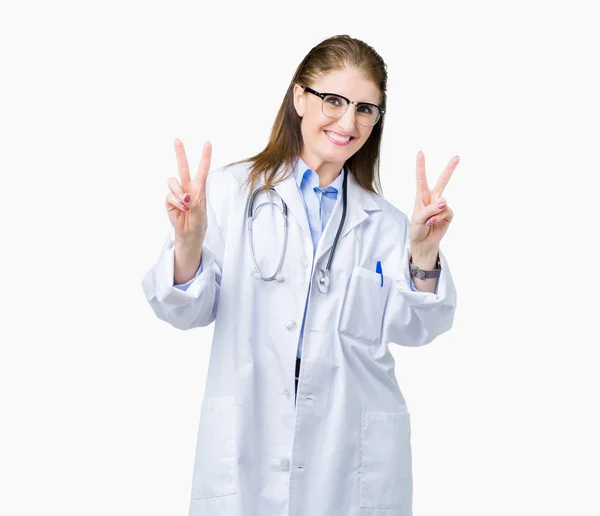 中年成熟的医生妇女穿着医疗外套在孤立的背景微笑着看着镜头显示手指做胜利的标志 — 图库照片