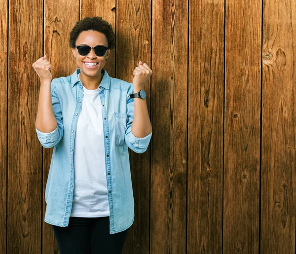 Güzel genç Afro-Amerikan kadın izole arka plan kutlama üzerinde güneş gözlüğü takıyor şaşırttı ve silah ile başarı için yükseltilmiş ve açık gözler hayrete düşürdü. Kazanan kavramı.