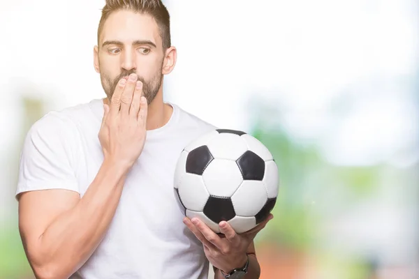 Young man holding soccer football ball over isolated background cover mouth with hand shocked with shame for mistake, expression of fear, scared in silence, secret concept