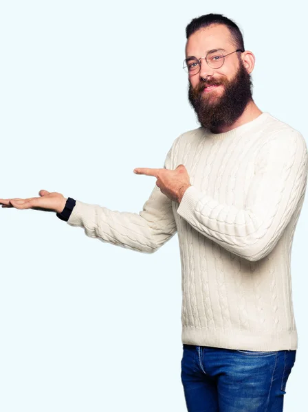 メガネと冬のセーターを着ている若いヒップスターの男性が 手でプレゼンしながら指で指さしながら カメラに驚嘆し 微笑んだ — ストック写真