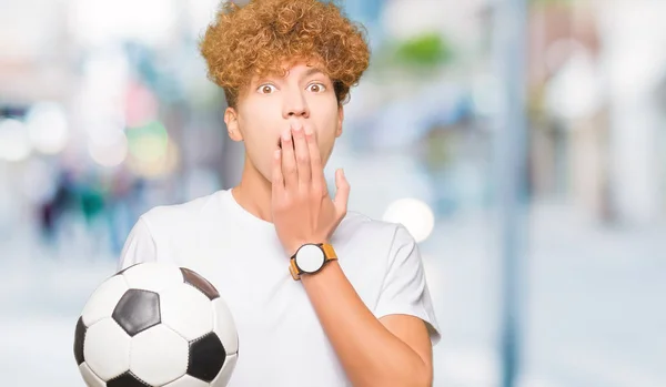 Young handsome man holding soccer football ball cover mouth with hand shocked with shame for mistake, expression of fear, scared in silence, secret concept