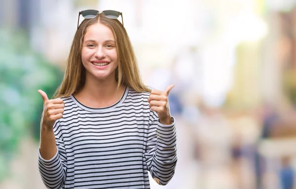 Young beautiful blonde woman wearing sunglasses over isolated background success sign doing positive gesture with hand, thumbs up smiling and happy. Looking at the camera with cheerful expression, winner gesture.