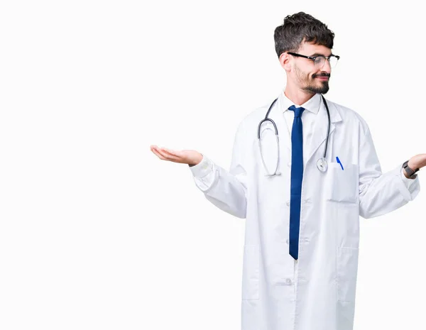 年轻的医生在孤立的背景上穿上医院外套微笑着露出双手张开手掌 呈现和广告比较和平衡 — 图库照片