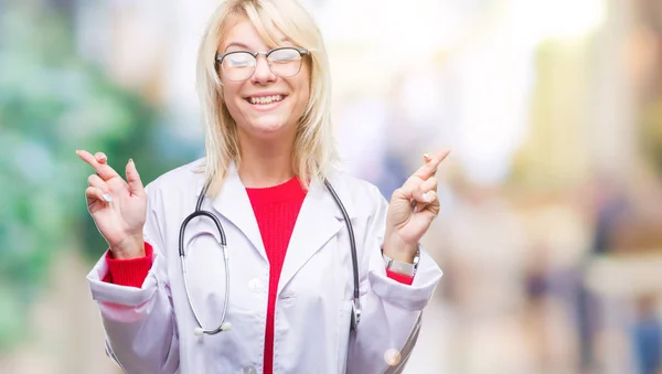 希望とは目を閉じて交差指の笑みを浮かべて隔離された背景の上医療制服を着て美しい金髪女医女性 運と迷信的な概念 — ストック写真