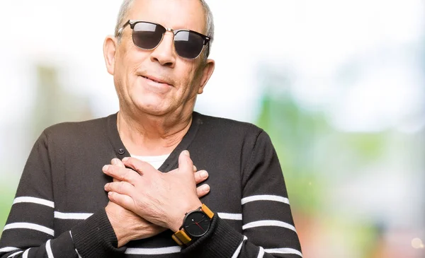 Handsome senior man wearing winter sweater and sunglasses smiling with hands on chest with closed eyes and grateful gesture on face. Health concept.