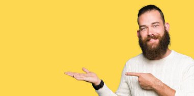 Genç hipster adam giyen kış kazak hayret ve el ile sunulması ve parmak ile işaret ederken kameraya gülümseyen.