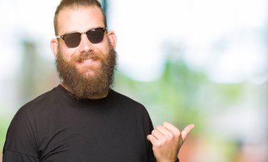 Genç hipster adam arıyor ve başparmak yanında yukarıyı mutlu yüz ile gülümseyen güneş gözlüğü takıyor.