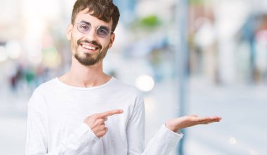 Yakışıklı delikanlı hayrete ve el ile sunulması ve parmak ile işaret ederken kameraya gülümseyen izole arka plan üzerinde güneş gözlüğü takıyor.