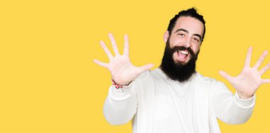 Uzun saç ve sakal gösterilen ve parmakları ile işaret sportif sweatshirt giyen genç adamla kendinden emin ve mutlu gülümseyerek süre on numara.