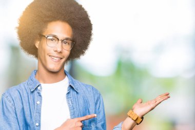 Hayret ve el ile sunulması ve parmak ile işaret ederken kameraya gülümseyen gözlüklü afro saçlı genç Afro-Amerikan adam.