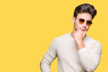 Genç yakışıklı adam çene düşünme hakkında soru, dalgın ifade üzerine el ile izole arka plan üzerinde güneş gözlüğü takıyor. Düşünceli yüzüyle gülümsüyor. Şüphe kavramı.