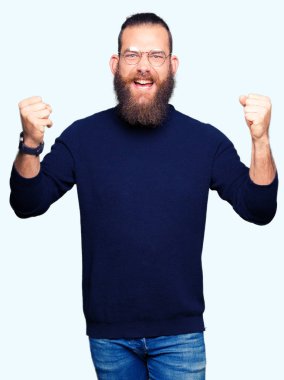 Gözlük ve balıkçı yaka kazak kutluyor giyen genç sarışın adam şaşırttı ve başarı için kaldırdı kollarını açarak hayret ve gözleri açık. Kazanan kavramı.