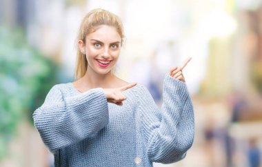 Gülümseyen ve iki elleri ve parmakları ile tarafına işaret kamera bakarak izole arka plan üzerinde kış kazak giyen genç güzel sarışın ve mavi gözlü kadın.