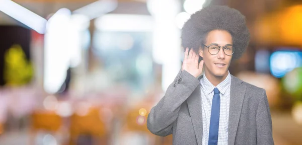 Young african american business man with afro hair wearing glasses smiling with hand over ear listening an hearing to rumor or gossip. Deafness concept.