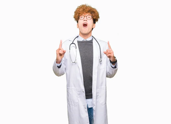 年轻英俊的医生男子穿着医疗外套惊讶和惊讶地抬起头来 用手指指指点点 举起双臂 — 图库照片