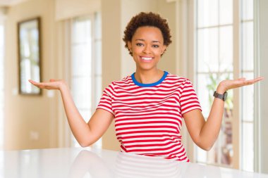 Genç ve güzel afro-amerikan kadın evinde gülümsüyor iki elini de açık gösteriyor, sunuyor ve reklam karşılaştırması yapıyor ve dengede duruyor.