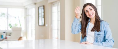 Evdeki beyaz masada oturan güzel genç bir kadının geniş açılı fotoğrafı. İtalyan jestleri yapıyor. Elleri ve parmakları kendinden emin ifade ediyor.