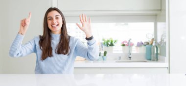 Evde beyaz masada oturan ve kendinden emin ve mutlu gülümserken parmaklarıyla işaret eden güzel genç kadının geniş açılı resmi.