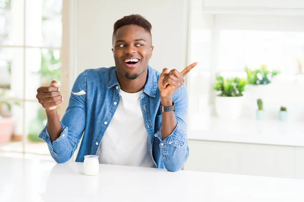 African american man eating healthy natural yogurt with a spoon very happy pointing with hand and finger to the side