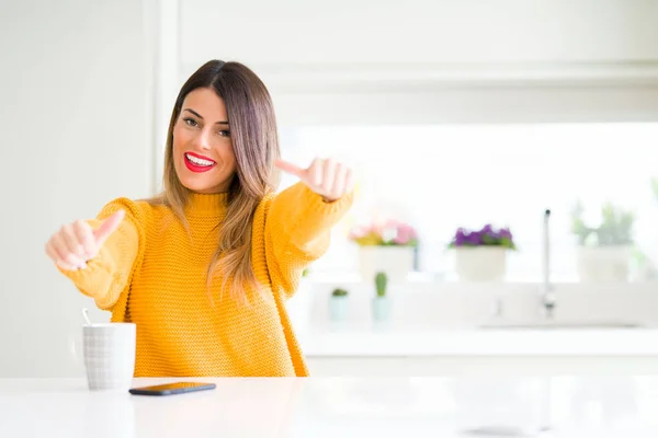 Young beautiful woman drinking a cup of coffee at home approving doing positive gesture with hand, thumbs up smiling and happy for success. Looking at the camera, winner gesture.