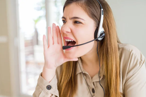 Beautiful young operator woman wearing headset at the office shouting and screaming loud to side with hand on mouth. Communication concept.