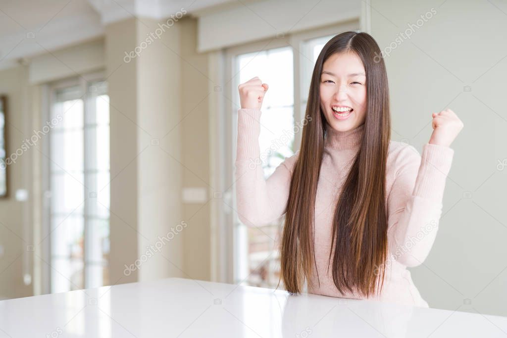 Beautiful Asian woman wearing casual sweater on white table very happy and excited doing winner gesture with arms raised, smiling and screaming for success. Celebration concept.