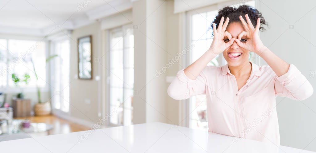 Wide angle of beautiful african american woman with afro hair doing ok gesture like binoculars sticking tongue out, eyes looking through fingers. Crazy expression.