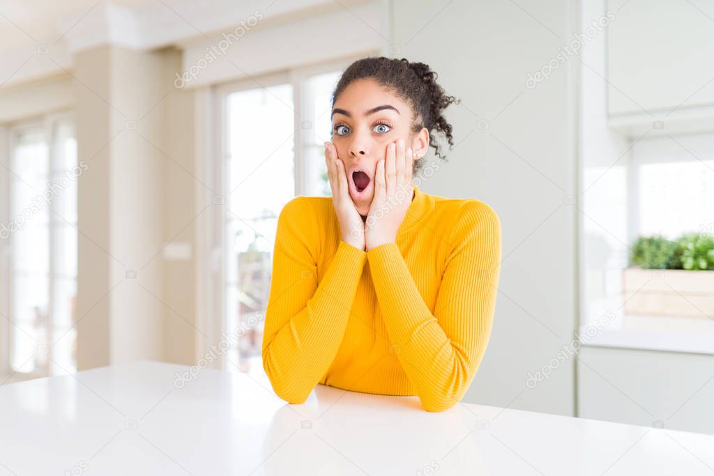 Beautiful african american woman with afro hair wearing a casual yellow sweater afraid and shocked, surprise and amazed expression with hands on face