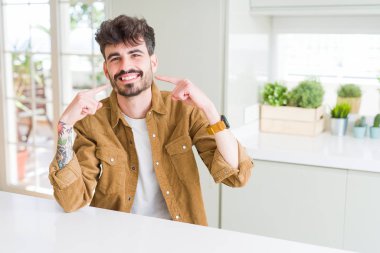 Beyaz masada oturan rahat ceket giyen genç adam emin gösteren gülümseyen ve parmaklar diş ve ağız ile işaret. Sağlık konsepti.