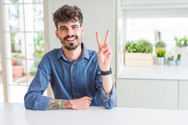 Genç adam, beyaz masanın üzerinde oturan rahat gömlek giyen kamera parmakları zafer işareti yapıyor gösteren bakıyor gülümseyen. İki numara.