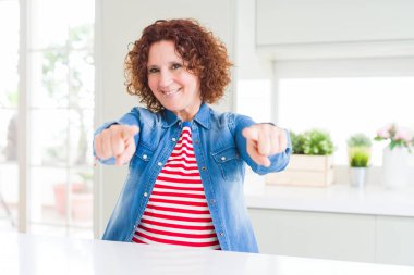 Evde kot ceket giyen kıvırcık saçlı orta yaş yaşlı kadın sizi ve parmaklarıile kamera işaret, olumlu ve neşeli gülümseyen