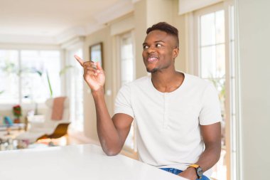 Yakışıklı afro-amerikan adam evde yüzünde büyük bir gülümseme ile beyaz masada, kameraya bakarak yan el ve parmak ile işaret.