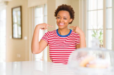 Genç güzel Afrikalı-Amerikalı kadın evde kendini gururlu ve mutlu parmakları ile işaret emin yüz, gülümseme ile seyir.