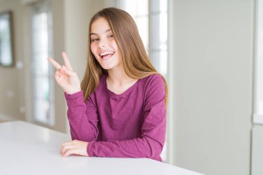 Beyaz masada güzel genç kız çocuk mutlu yüz zafer işareti yapıyor kamera göz kırpıyor ile gülümsüyor. İki numara.